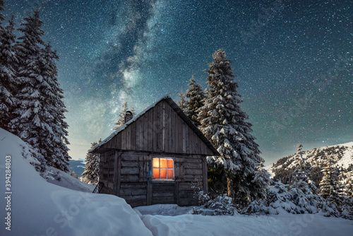 Fantastic winter landscape with wooden house in snowy mountains. Starry sky with Milky Way and snow covered hut. Christmas holiday and winter vacations concept