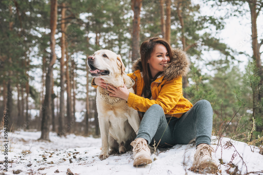 Young smiling woman in yellow jacket with big kind white dog Labrador walking in the winter forest Young smiling woman in yellow jacket with big kind white dog Labrador walking in the winter forest