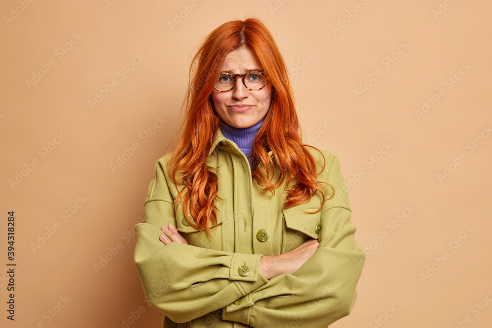 Offended dissatisfied redhead young woman keeps arms folded waits for explanations from boyfriend has hurt feelings dressed in fashionable green jacket smirks face poses against brown background