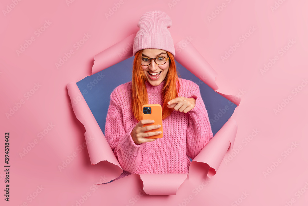 Cheerful surprised redhead woman points at smartphone screen reacts on big discounts in online store laughs happily and looks at something unbelievable wears stylish clothes poses through torn hole Cheerful surprised redhead woman points at smartphone screen reacts on big discounts in online store laughs happily and looks at something unbelievable wears stylish clothes poses through torn hole