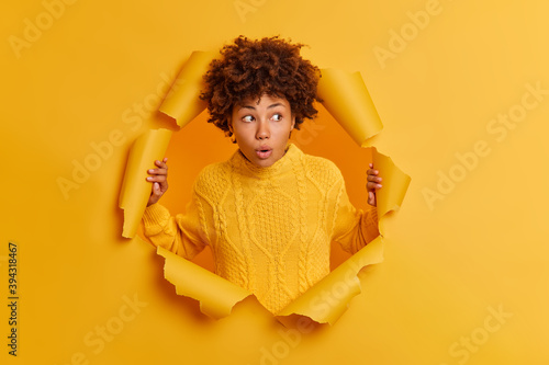 Embarrassed shocked young Afro American woman stares with bugged eyes, keeps mouth opened, wears sweater, reacts on shocking news breaks through paper background. People and emotions concept photo