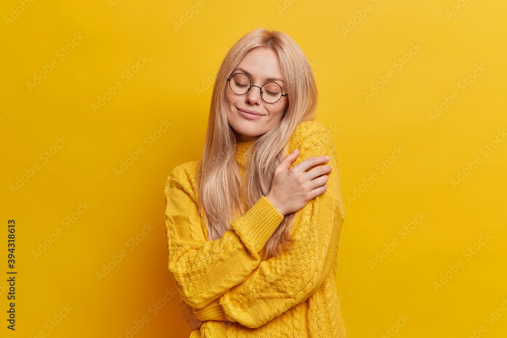 Selfish emotion concept. Pleased relaxed blonde European woman hugs herself gently expresses self love and happiness closes eyes wears warm knitted sweater isolated over vibrant yellow background. Selfish emotion concept. Pleased relaxed blonde European woman hugs herself gently expresses self love and happiness closes eyes wears warm knitted sweater isolated over vibrant yellow background.