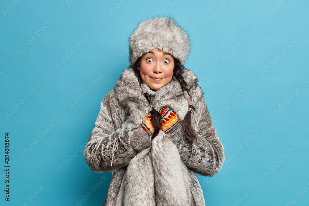 Winter and Christmas concept. Surprised Asian woman with two pigtails dressed in winter fur hat coat shocked to see heavy snowfall outside poses against blue background. Arctic female in warm clothing Winter and Christmas concept. Surprised Asian woman with two pigtails dressed in winter fur hat coat shocked to see heavy snowfall outside poses against blue background. Arctic female in warm clothing