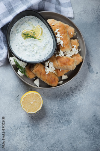 Filo mini rolls with feta cheese stuffing and tzatziki dip, above view on a light-blue stone background, vertical shot with copy space