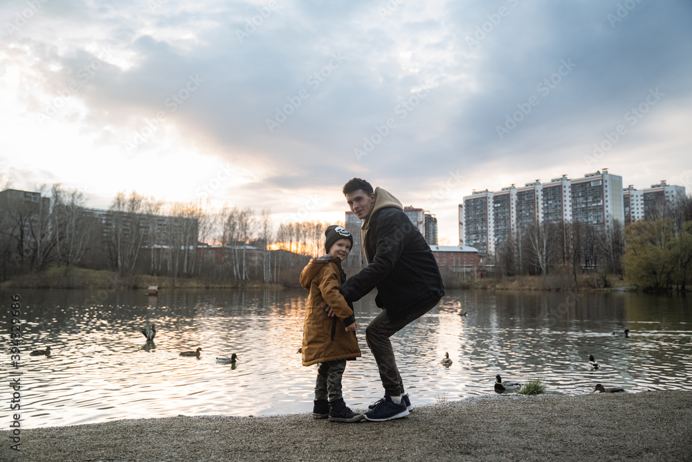 looking at the camera, Silhouettes of a father and son standing by the lake, ducks swim, the sun goes down, one of the parents with a child is walking against the background of a high-rise building, t
