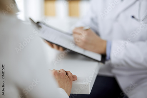 Unknown male doctor and patient woman discussing current health examination while sitting in clinic and using clipboard. Perfect medical service in hospital. Medicine and healthcare concept © cameravit