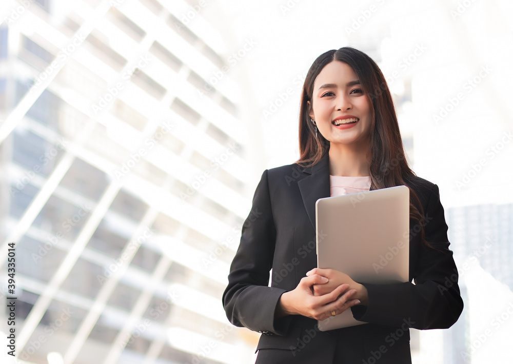 Young Asian business woman wearing black suit, holding computer laptop, smiling and looking at camera. Outdoor with business building background. Young Asian business woman wearing black suit, holding computer laptop, smiling and looking at camera. Outdoor with business building background.