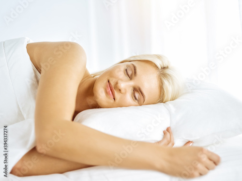 Beautiful young woman sleeping while lying in bed comfortably and blissfully in sunny bedroom. Good morning concept