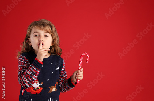 Cute little girl with sweet Christmas candy cane showing silence gesture against red background