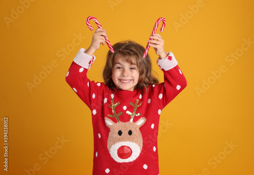 Cute little girl in Christmas sweater holding sweet candy canes near head against orange background