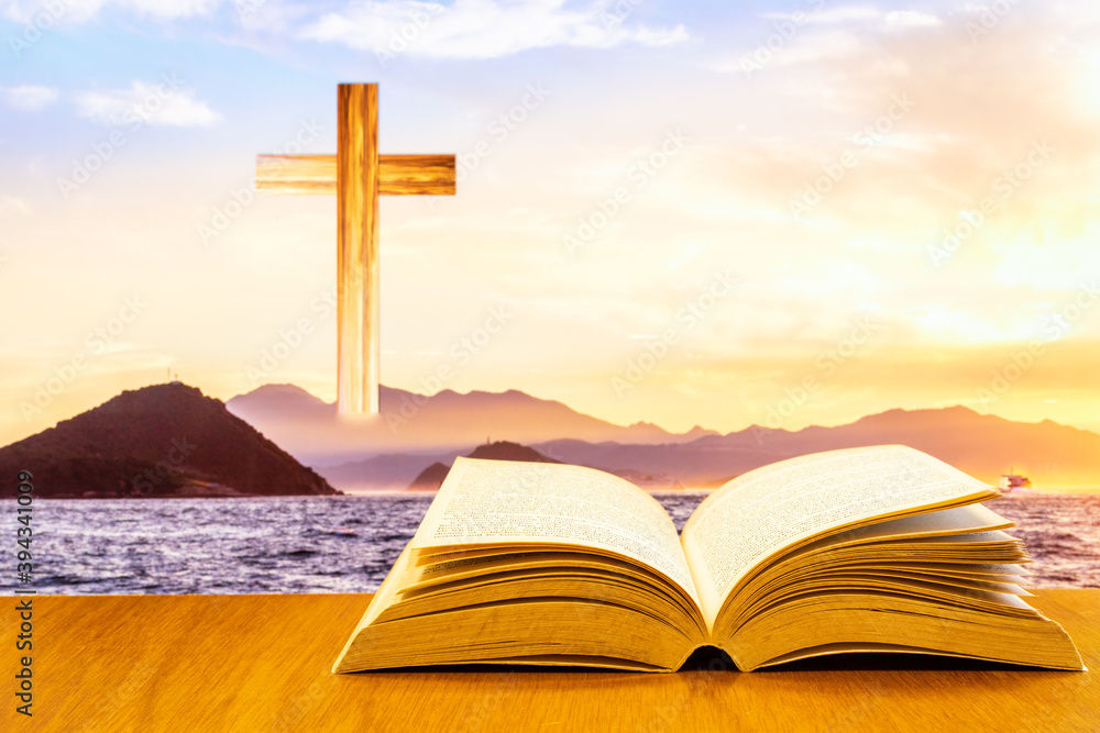 Open old bible on a wood table with blurred cross & colourful clouds and sky as background Open old bible on a wood table with blurred cross & colourful clouds and sky as background