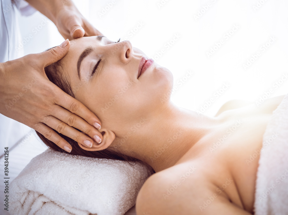 Beautiful brunette woman enjoying facial massage with closed eyes in sunny spa center . Relaxing treatment and cosmetic medicine concepts Beautiful brunette woman enjoying facial massage with closed eyes in sunny spa center . Relaxing treatment and cosmetic medicine concepts