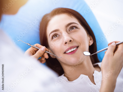 Smiling brunette woman being examined by dentist at sunny dental clinic. Hands of a doctor holding dental instruments near patient s mouth. Healthy teeth and medicine concept
