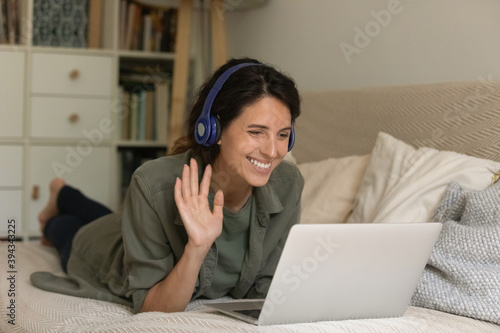 Smiling millennial female in headphones lying on couch at home wave greet talk on video call on laptop. Happy young woman in earphones have webcam digital conference or online web lesson on computer.