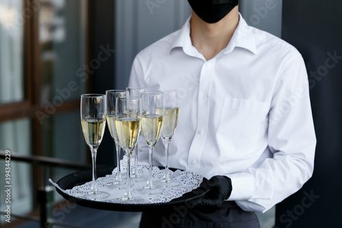 The waiter holds a tray with glasses of champagne. A waiter in a medical mask. Restaurant work during the coronavirus pandemic.