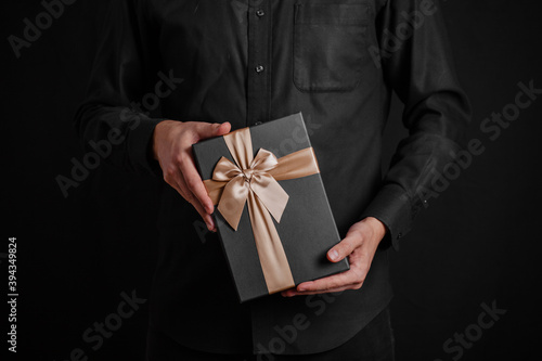A guy in a black shirt is holding a gift with a golden ribbon. Black background. Black gift box in hands..