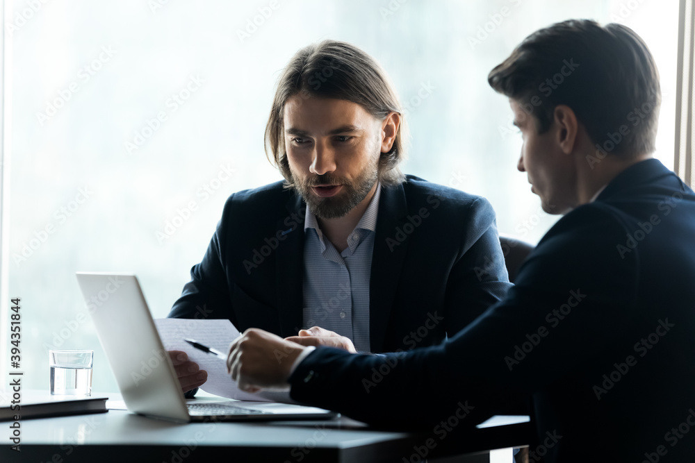 Focused Caucasian businessmen work on laptop at office meeting brainstorm considering financial paperwork. Serious male business partners work on computer talk discuss cooperation at briefing. Focused Caucasian businessmen work on laptop at office meeting brainstorm considering financial paperwork. Serious male business partners work on computer talk discuss cooperation at briefing.