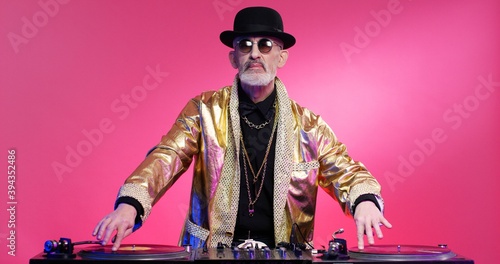 Vintage stylish fancy retired man in hat and sparkling golden coat playing music as DJ on special mixer at pink wall background. Retro disco concept. Grandpa working dj at party.