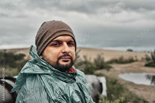 A man with a beard in a green raincoat and hat, face close-up. Overcast weather.