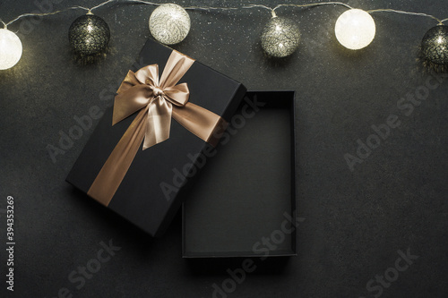 Opened black gift box with golden ribbon. Gift on a black granite surface. Christmas lights above the box..