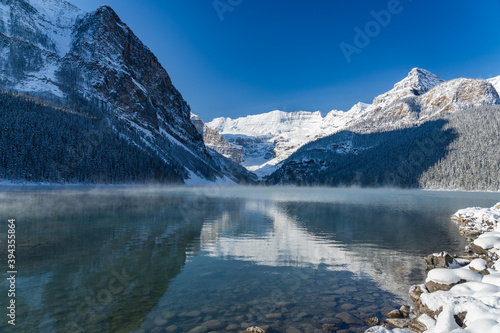 Lake Louise in early winter sunny day morning. Mist floating on turquoise color water surface. Clear blue sky, snow capped mountains in background. Beautiful natural landscape in Banff National Park.