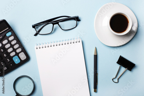 Calculator, blank notebook, pen, coffee cup, magnifier and glasses on a light blue background. Top view close up