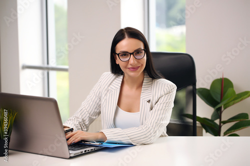 Cheerful brunette lady with important document friendly smiling and looking at camera while browsing laptop at desk in home office