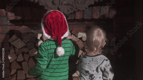 Two little brother boys in santa cap standing by fireplace at xmas eve. Kids clebrating merry christmas and happy new year at home. Cute children standing back together at night photo