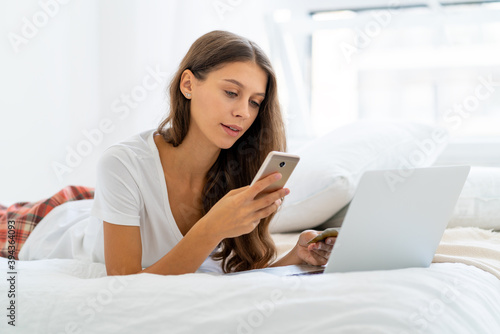 Young woman shopping online, entering code from SMS message on mobile phone, security. Female sitting on bed in bedroom and working from home on laptop