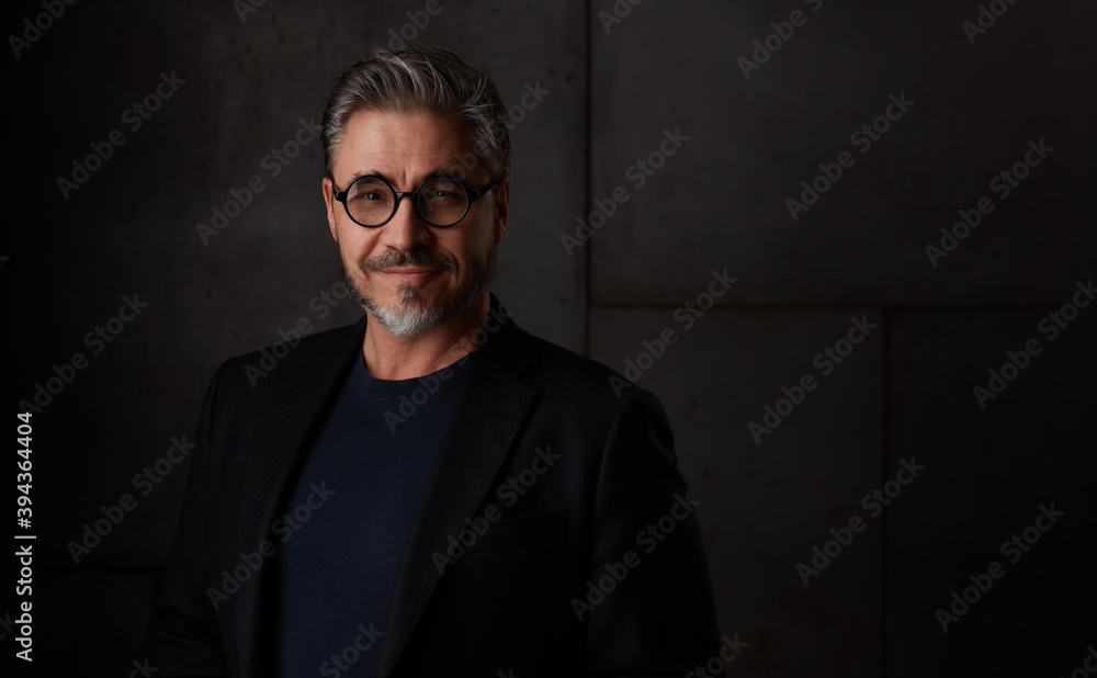 Obraz premium Portrait of happy older man in 50s with gray hair and glasses against dark background. Wearing jacket, smiling, copy space.