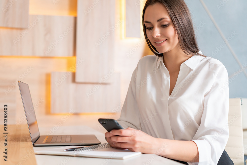 Office manager woman in white shirt, sitting to table with phone in hands and notebook with laptop on the table. Smiling office worker in big light office room. Concept of work Office manager woman in white shirt, sitting to table with phone in hands and notebook with laptop on the table. Smiling office worker in big light office room. Concept of work