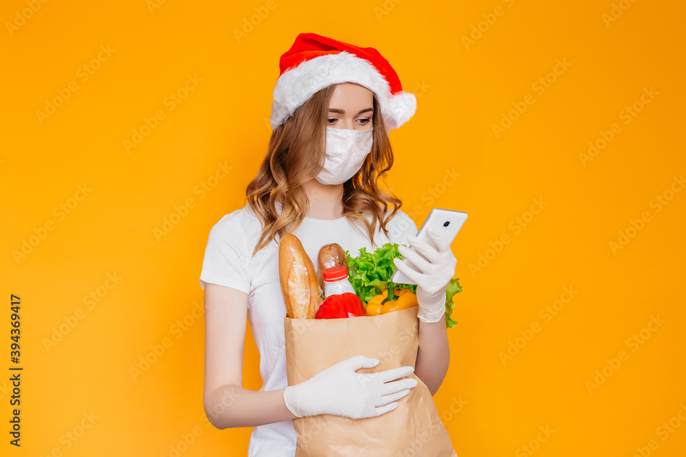 New Year delivery. Young woman courier wearing a medical mask, gloves looks at a mobile phone and holds an eco paper bag with food, vegetables. Safe delivery service, coronovirus, vegan eco food New Year delivery. Young woman courier wearing a medical mask, gloves looks at a mobile phone and holds an eco paper bag with food, vegetables. Safe delivery service, coronovirus, vegan eco food