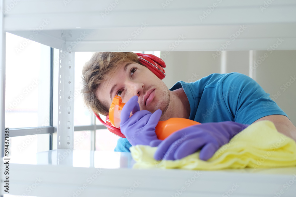 Young Caucasian male Wearing violet rubber gloves is holding Cleanser bottle and sponge. He is cleaning and wiping on white Shelf for housework.Lifestyle of Familyman on Holiday Concept. Young Caucasian male Wearing violet rubber gloves is holding Cleanser bottle and sponge. He is cleaning and wiping on white Shelf for housework.Lifestyle of Familyman on Holiday Concept.