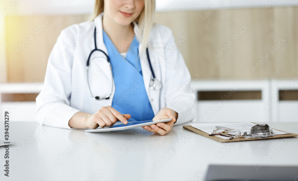 Unknown woman-doctor sitting in sunny clinic and using tablet pc, close-up. Data and best service in medicine Unknown woman-doctor sitting in sunny clinic and using tablet pc, close-up. Data and best service in medicine