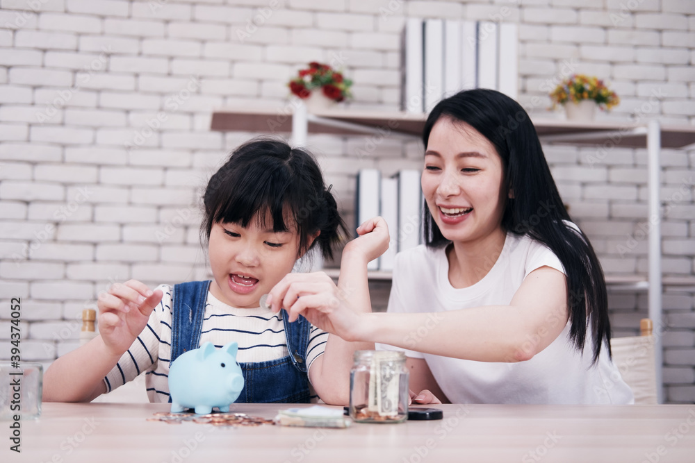 Fototapeta premium Smiling Asian little asian girl child is putting coins into piggy bank for saving money for the future with mother on wooden table. Child educational for homeschool concept.