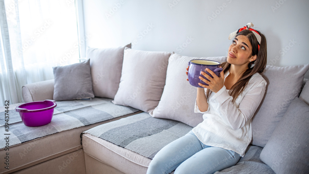 Obraz premium Frustrated homeowner watching ceiling leaks sitting on a couch at home. Worried Woman Holding Two Buckets While Water Droplets Leaking From Ceiling.