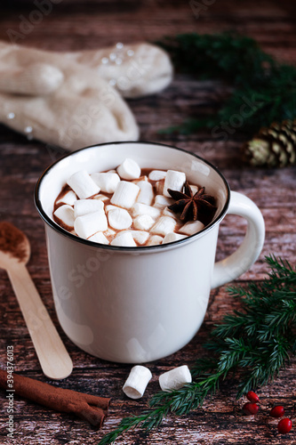 White cup of hot spiced cacao or chocolate with marshmallows on a rustic wooden tabletop with fir branches and knitted gloves in the background. 