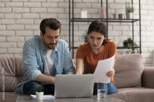 Focused young married couple looking at computer screen, involved in planning monthly household budget, holding paper bills making payments online in banking application, doing financial paperwork.