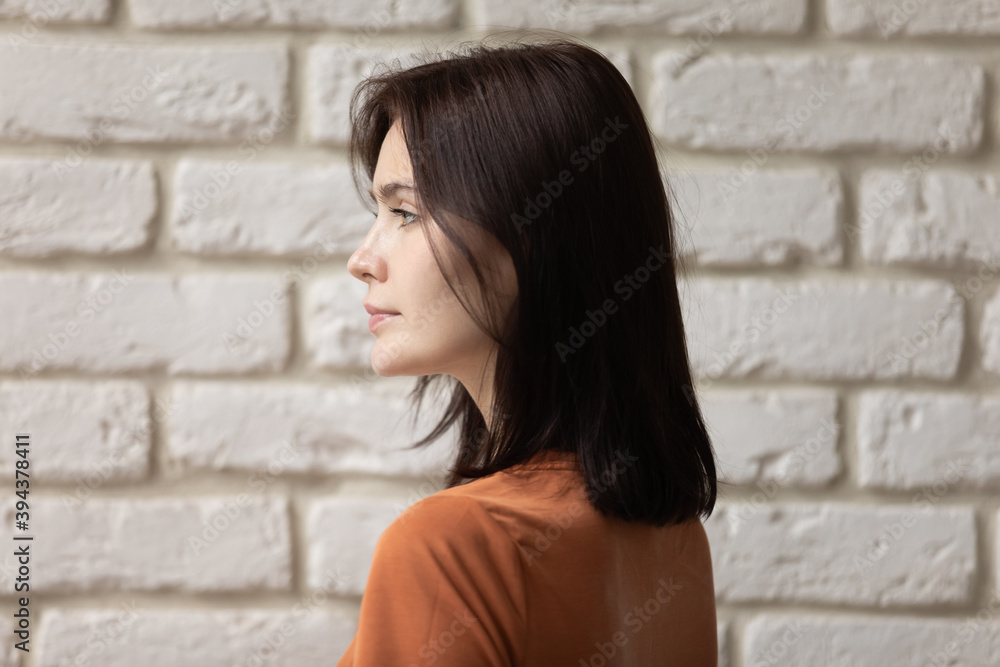 Side head shot pretty millennial brunette woman with shoulder length hair looking away, standing near brick wall, satisfied with hairdresser coloring work, dreamy lady visualizing planning future.