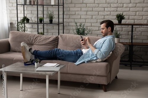 Full length young 30s man in casual wear lying on comfortable sofa, using mobile software applications, web surfing information, shopping goods or services online, technology addiction concept.