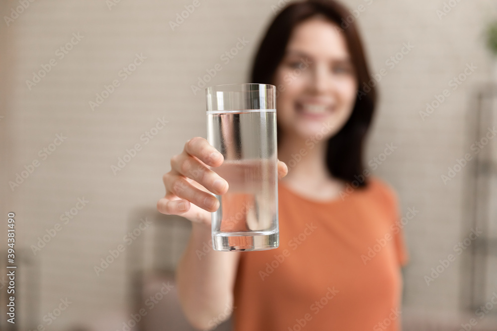 Close up focus on glass with pure mineral clear water in female hands, happy young woman reaching out hand to camera, offering drinking filtered aqua every day, daily healthcare morning habit. Close up focus on glass with pure mineral clear water in female hands, happy young woman reaching out hand to camera, offering drinking filtered aqua every day, daily healthcare morning habit.