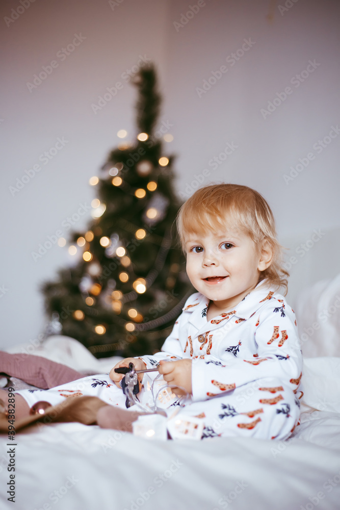 the child's first Christmas. a pensive little boy is lying in his holiday pyjamas on a bed against the background of a brightly decorated fir tree with a Golden light garland. the child's first Christmas. a pensive little boy is lying in his holiday pyjamas on a bed against the background of a brightly decorated fir tree with a Golden light garland.