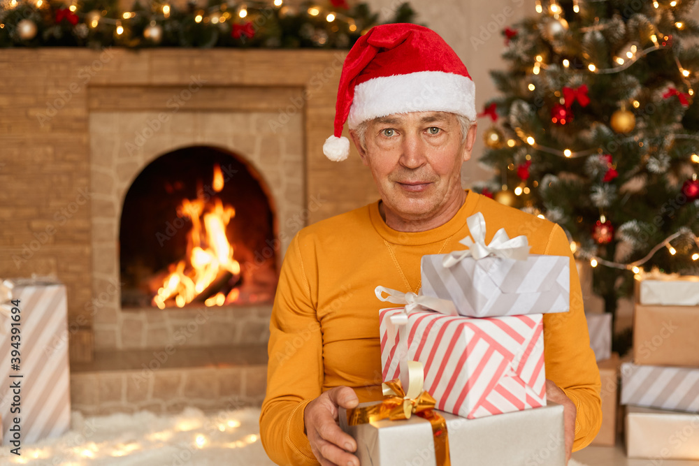 Senior wrinkled man sitting in living room with Christmas decoration and holding stack of present boxes, looking ar camera, posing near fireplace and xmas tree. Senior wrinkled man sitting in living room with Christmas decoration and holding stack of present boxes, looking ar camera, posing near fireplace and xmas tree.