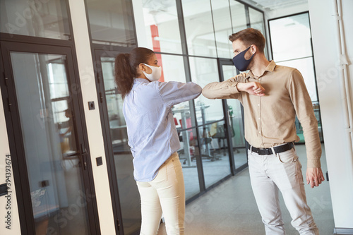 Covid 19 infection prevention. Two young diverse colleagues wearing face protective masks bumping elbows, greeting each other while standing in the modern office