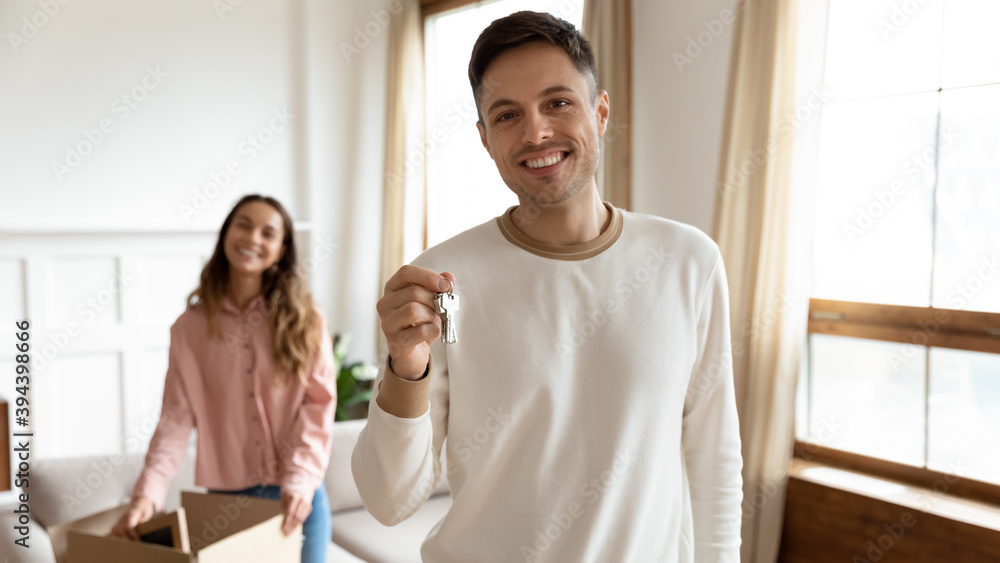 Obraz premium Excited millennial man smiling looking at camera showing keys to the new first house, on background happy wife holding cardboard box unbox belongings. Bank loan, mortgage, real-estate buyers concept
