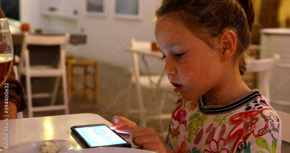 Portrait of six years old cute child with white dress, playing piano in smartphone display, sitting in a white table dinning in a restaurant next to her mother
Portrait of six years old cute child with white dress, playing piano in smartphone display, sitting in a white table dinning in a restaurant next to her mother