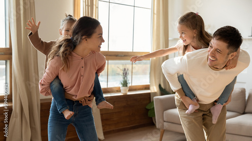 Cheerful parents playing with two preschool adorable daughters enjoy active games in living room piggyback riding children running laughing feels happy. Leisure activity with kids on weekend concept