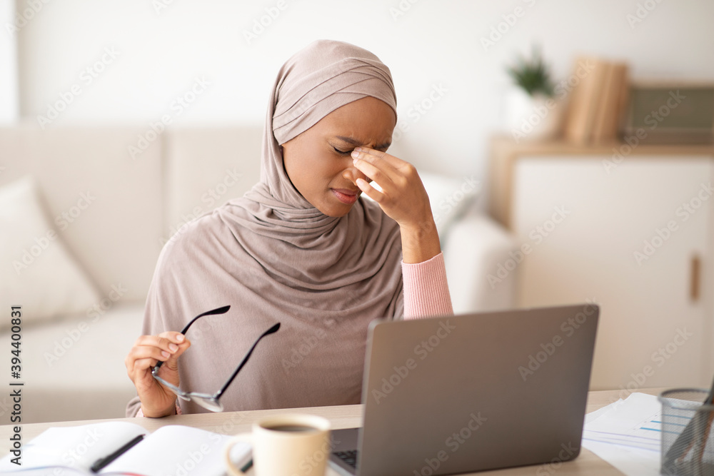 Tired black woman in hijab rubbing her eyes while working online on laptop at home Tired black woman in hijab rubbing her eyes while working online on laptop at home