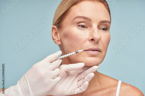 Attractive mature caucasian woman making lip augmentation procedure by cosmetologist while standing against blue background
