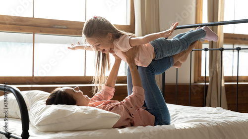 Billede på lærred In the morning woman lies in bed play with 6s daughter, hold cute kid, girl stretched arms like airplane wings flying in air have fun with mom